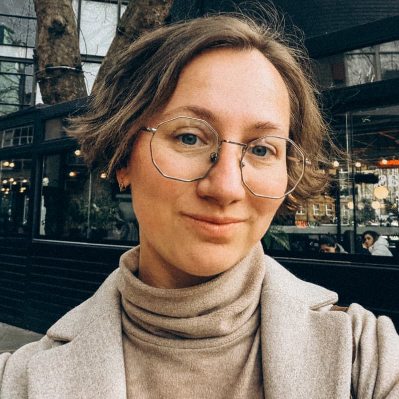 Alina Kabanets, smiling, in round glasses and a camel coat on a London street